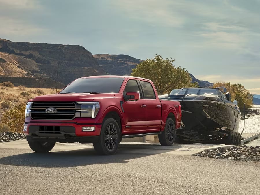 Red Ford truck towing a black boat on a scenic road, surrounded by mountains and trees.
