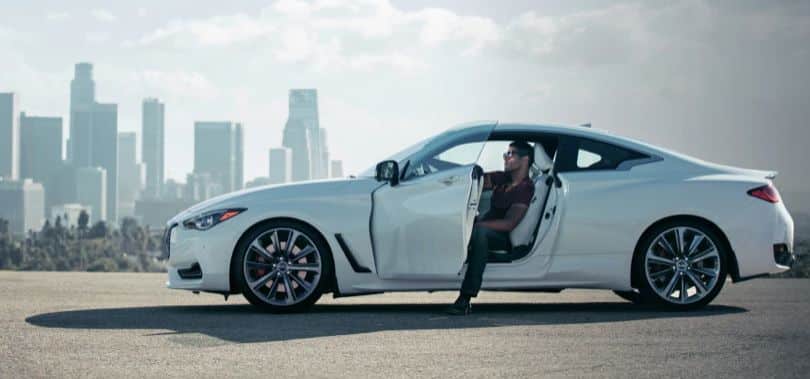 A person sits in a white sports car with the door open, set against a city skyline.